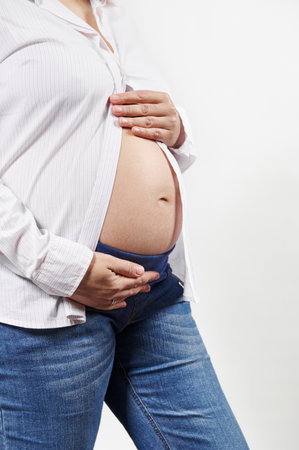 Midsection of a pregnant woman, expecting baby, enjoying her 28 week of pregnancy, holding hands on her bare belly, isolated on white background. Close-up of gravid female. Maternity. Motherhood.の写真素材