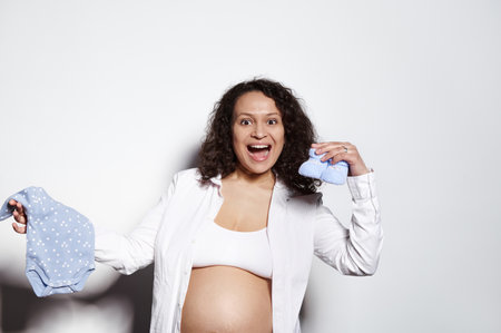 Overjoyed emotional multi ethnic curly haired pregnant woman holding baby clothes, blue bodysuit and booties for newborn boy, smiles looking at camera, posing bare belly over isolated white backgroundの写真素材