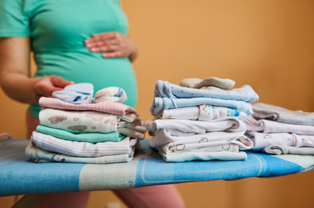 A stack of folded ironed clean baby clothes on ironing board, against the background of blurred pregnant woman in turquoise t-shirt, gently caressing her belly. Pregnancy. Preparation to childbirthの写真素材