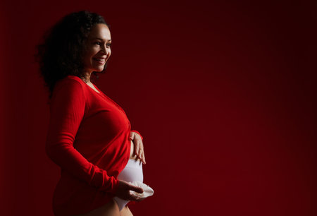 Delightful pregnant woman dressed in red shirt, holding a white orchid flower, smiling cutely, caressing her big belly, expressing positive emotions, feeling baby movements, isolated on red backgroundの写真素材