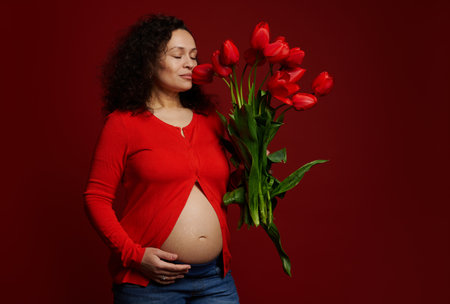 Beautiful curly Latin American pregnant woman, sniffing cute red tulips, touching her belly with stretch marks from pregnancy, posing with her eyes closed on isolated red background. Body positivityの写真素材