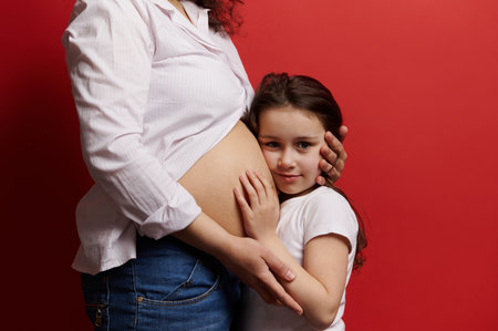 Beautiful little girl hugging the belly of her pregnant mother, looking at camera, isolated on red background. Gravid woman with naked tummy embracing her lovely daughter. Pregnancy. Family. Maternityの写真素材