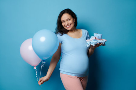 Multi-ethnic smiling happy pregnant woman with big belly, holding newborn clothes and two balloons...