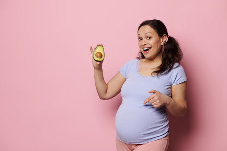 Studio shot of smiling young vegetarian expectant woman, holding one half of fresh avocado fruit, pointing at her pregnant belly over isolated pink background. Healthy nutrition and pregnancy conceptの写真素材