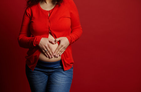 Close-up pregnant woman in casual clothes, makes heart shape with her hands over her bare belly, isolated on red background. Happy carefree pregnancy concept. Enjoying maternity leave. Womens healthの写真素材