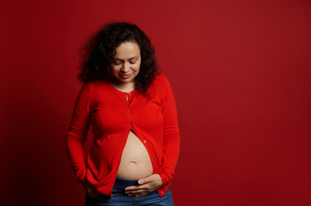 African American positive adult pregnant woman, touching her belly, smiling, enjoying feeling baby movements, isolated on red background with copy advertising space. Pregnancy 28 week. Expecting babyの写真素材