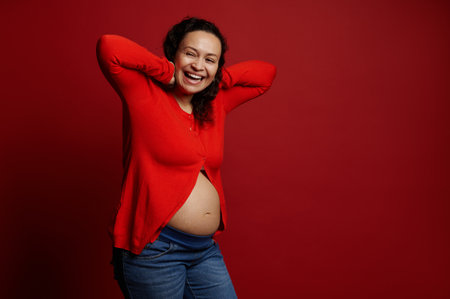 Curly haired ethnic pregnant adult woman laughing, posing bare belly over isolated red background. Enjoying wonderful moments of happy carefree pregnancy. Copy space. Maternity. People. Lifestyleの写真素材