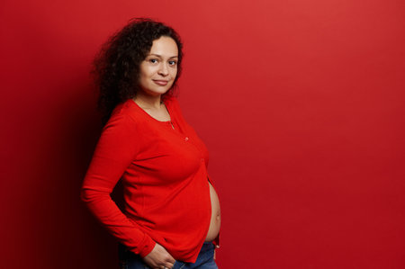 Confident portrait of a dark haired curly beautiful pregnant woman in red shirt, smiling looking at camera, isolated on red background. Copy advertising space. Pregnancy. Maternity. Body positivity.の写真素材