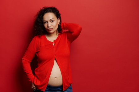 Confident studio portrait of attractive curly multi ethnic brunette, beautiful pregnant woman, expectant mother, looking at camera, dressed in red shirt, shows her bare belly, isolated red backgroundの写真素材