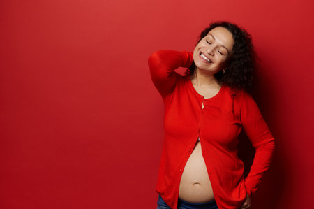 Authentic studio portrait of a middle aged confident beautiful pregnant woman, expectant mother posing bare belly and closed eyes, smiling over isolated red color background. Pregnancy. Maternityの写真素材