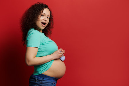 Attractive sexy pretty woman, pregnant expectant mother looking at camera, posing on red color backdrop with cute baby booties on her bare belly. Happy carefree pregnancy and maternity. Expecting babyの写真素材
