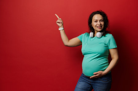 Multi ethnic dark-haired positive pregnant woman in casual blue turquoise t-shirt and jeans, smiling looking at camera, pointing her index finger aside a copy space over red isolated studio backgroundの写真素材