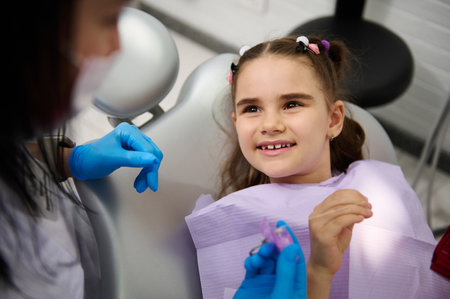 Multi ethnic little kid girl in dentists chair, smiles broadly to her doctor dentist hygienist during regular dental check-up at pediatric at dental clinic. Pediatric dentistry. Child teeth therapyの写真素材
