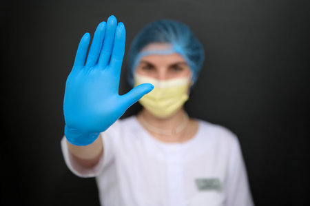 Details on the hand in blue medical glove of female doctor, medic, healthcare worker showing stop sign with hand, dressed in white medical gown and yellow surgical mask, isolated on black backgroundの写真素材