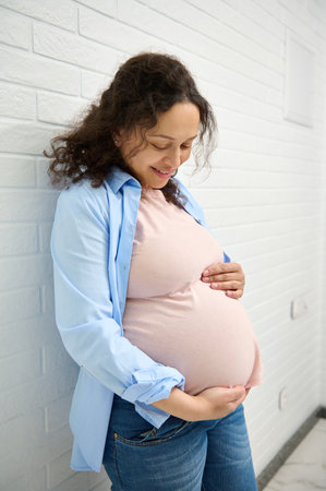 Confident portrait of a multi ethnic beautiful pregnant woman in 30 week of pregnancy,gorgeous gravid expectant mother smiling and gentry stroking her belly, standing over white brick wall background.の写真素材