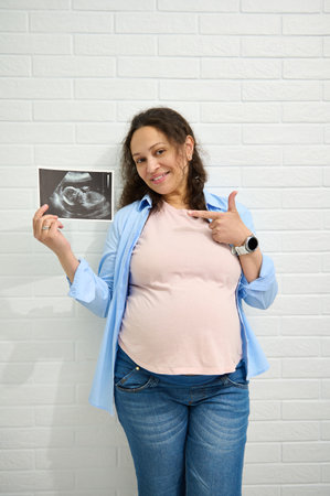 Happy middle aged pregnant woman, expectant mother, smiling, looking at the camera, pointing aside at the ultrasound of her baby, isolated against white brick wall background. Motherhood. Pregnancyの写真素材