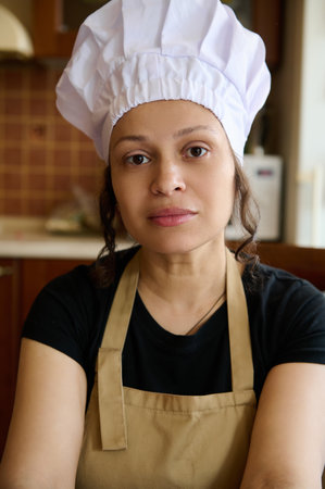 Confident portrait of a beautiful multi ethnic woman housewife, female chef baker confectioner in white hat and beige apron, confidently looking at camera. People. Profession. Career. Hobby, Lifestyleの写真素材
