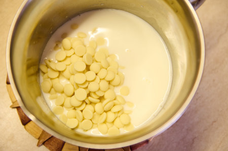 Top view of white organic belgian chocolate chips inside a pan with hot milk on kitchen worktop. Melting Chocolate. Preparing delicious cream for soaking sponge biscuits or a glaze for decorating cakeの写真素材