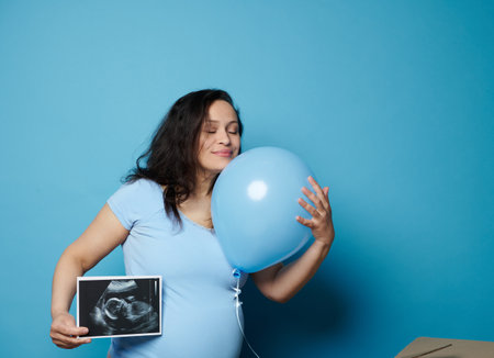 Delightful conscious pregnant woman gently hugging a blue balloon, posing with ultrasonography image of her future baby boy, experiencing happy emotions of her pregnancy, isolated over blue backgroundの写真素材