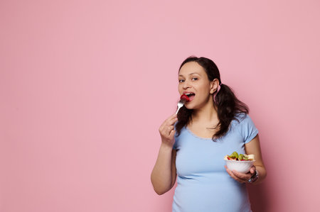 Isolated portrait on pink background of a glamour charming pregnant woman in blue t-shirt eating...