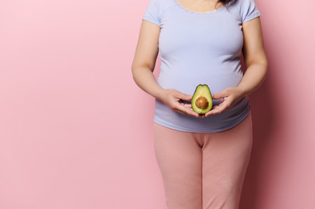 Midsection of pregnant expectant woman holding a half of avocado fruit at the level of her big belly, isolated over pink background. Pregnancy. Prenatal and maternity lifestyle. Womens health conceptの写真素材