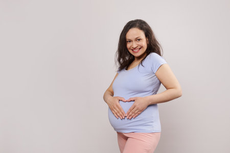 Beautiful happy pregnant woman holding hands on her belly, forming a heart shape, smiling looking at camera, isolated on white background with copy space. Happy pregnancy. Maternity. Expecting a babyの写真素材