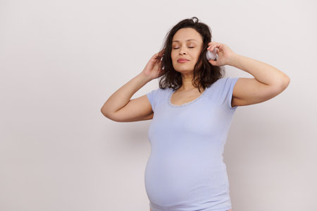 Multi ethnic brunette, happy adult pregnant woman listening to soothing music on wireless headphones, posing with her eyes closed over white isolated background. Happy pregnancy. Maternity conceptの写真素材