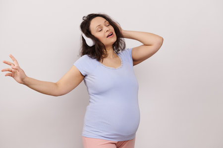Multi ethnic cheerful delightful pregnant woman in blue t-shirt, singing and dancing while listening to soothing music on wireless headphones, posing with her eyes closed on white isolated backgroundの写真素材