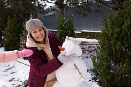 Multi ethnic cheerful woman in warm winter clothes, smiles looking at camera, having fun while making a snowman in the snow covered backyard. Winter holidays. People. Leisure activity. Christmastimeの写真素材