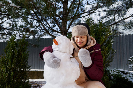 Happy cheerful positive woman making a snowman in the snow covered backyard, putting a carrot and tree branch on a huge snowball, enjoying happy winter weekend on a snowy cool february day. Wintertimeの写真素材