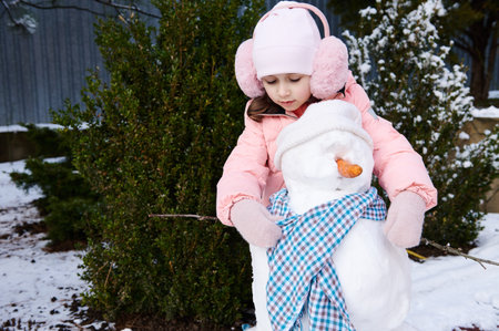 Charming cute little child girl puts on a blue shawl on a snowman, during active winter leisure games in the snowy backyard in the fresh air. Happy childhood and healthy lifestyle. Christmas timeの写真素材
