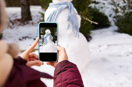 Close-up of hands holding smart mobile phone in live view mode, photographing snowman with blue shawl and carrot nose, in a snow covered nature background. Wintertime. Christmas time. Winter leisuresの写真素材