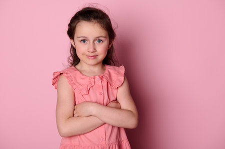 Lovely child girl wearing pink dress, standing with her arms folded over pink isolated background, smiles looking at camera. Happy carefree childhood concept. Kids fashion and beauty. Daughter. Familyの写真素材