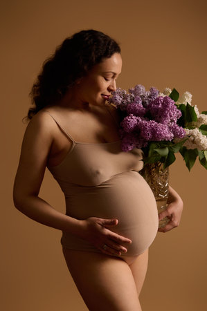 Attractive pregnant woman in beige underwear, posing with beautiful bunch of lilacs, isolated on beige background. Fashion portrait of authentic young gravid woman expecting a baby. Pregnancy 30 weeksの写真素材