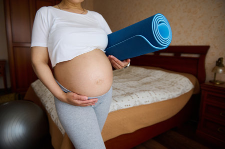 Midsection of a pregnant expectant woman mother holding her belly and fitness mat, standing at home bedroom. Prenatal stretching exercises. Sport. Workout. Yoga practice in pregnancy time. Maternityの写真素材