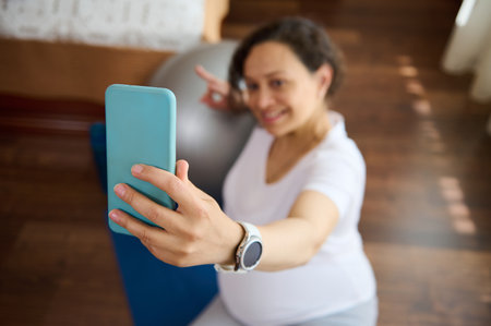 Close-up smart mobile phone in the hands of pregnant woman, expectant gravid mother taking selfie sitting on an exercise mat after prenatal fitness workout at home. Active healthy pregnancy lifestyleの写真素材