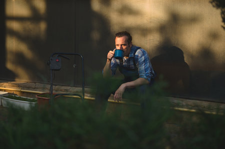 View through hedge on the foreground to professional male gardener drinking coffee at sunset, taking break after hard working day in the garden. People and Labour concept. Gardening. Landscaping themeの写真素材