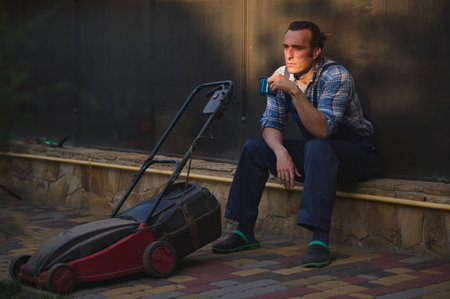 Sunset sun rays fall on face of a gardener with lawn mower, overworked after hard days work in the...