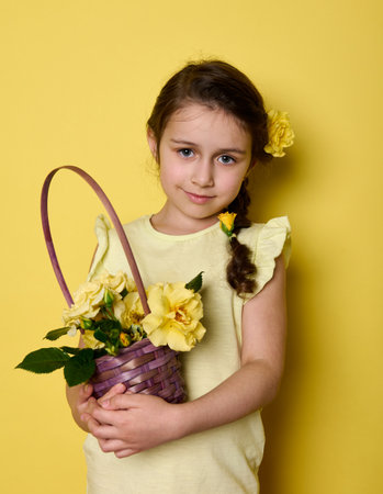 Beauty portrait Caucasian noble charming little girl in yellow elegant wear and flowers in hair, holding bouquet of yellow roses in purple wicker basket, smiling looking at camera, isolated backgroundの写真素材