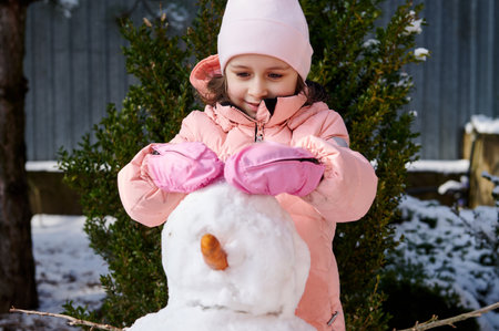 Authentic portrait of adorable little child girl playing outdoors winter games, building a big snowman in the backyard, smiling, wearing warm pink down jacket. Wintertime. Christmas holidays conceptの写真素材