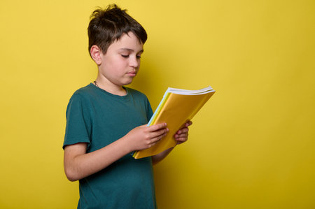 Adorable Caucasian schoolboy in green casual t-shirt, primary school student, holds colorful textbooks, isolated on yellow background. Back to school. Education concept. Smart clever kidsの写真素材