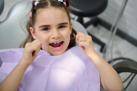 Close-up portrait of beautiful little girl using dental floss, brushing her teeth, sitting in the dentist's chair and smiling cutely, looking at the camera. Oral hygiene concept for caries preventionの写真素材