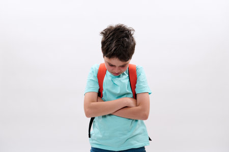 Offended distraught schoolboy with backpack, folding arms, standing tearful and looking down after bullying act at school, isolated on white background. Social issues. Problems. Puberty difficult ageの写真素材