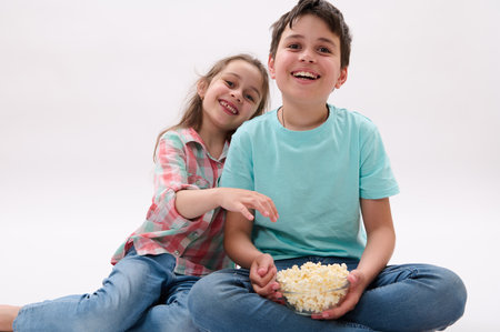 Caucasian amazed funny children, school kids, handsome teen boy and lovely little girl, brother and sister, having fun together, smiling, watching cartoons and eating popcorn. Lifestyle. Entertainmentの写真素材