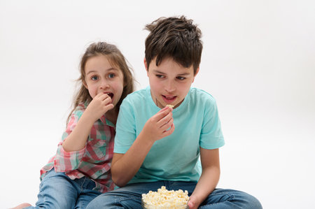 Caucasian adorable cheerful happy kids, preteen boy and little girl eating popcorn, watching movie or cartoons, smiling looking toward the camera, sitting together on isolated white studio backgroundの写真素材