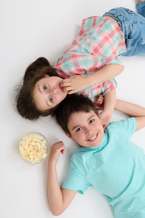 Top view happy kids, pre teen boy and little girl, brother and sister smiling looking at camera, lying on their backs on white studio background near a bowl with tasty popcorn, expressing positivityの写真素材
