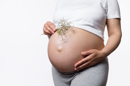 Close-up pregnant abdomen of gravid woman holding gypsophila bunch and embracing her big belly in...