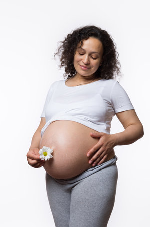 Charming smiling happy Latina gravid mother caressing her big pregnant belly in late pregnancy, holding daisy flower, isolated on white background. Women's health. Obstetrics and gynecology. Maternityの写真素材
