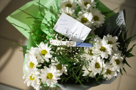 View from above of a beautiful bouquet of white chamomile flowers with greeting card and positive pregnancy test kit in the floral design studio. Happy Mothers Day. Arrangements and gifts. Copy spaceの写真素材