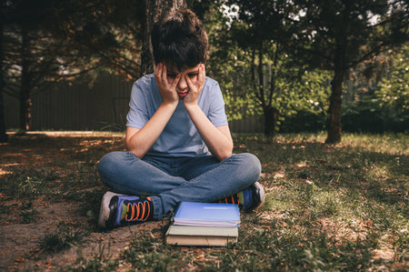 A lonely schoolboy teenager sitting sad and tired in a park under a tree with textbooks laid out, tired after the first days at school. Learning problems, adolescence, peer difficulties. Bullyingの写真素材